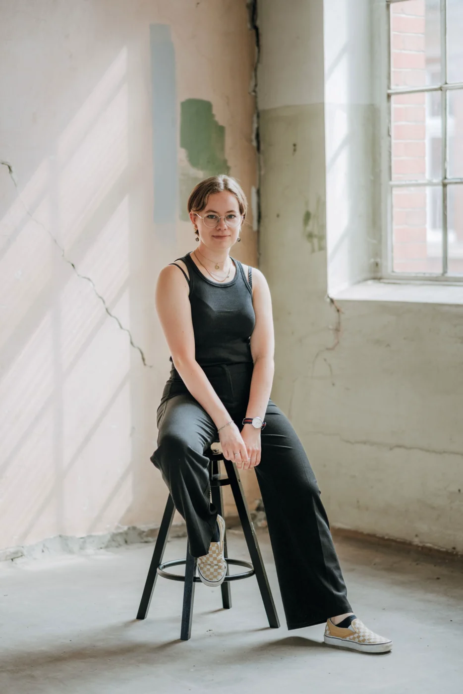 Eine Person in schwarzem Outfit sitzt auf einem Hocker in einem hellen Raum mit großen Fenstern und abblätternden Wänden.