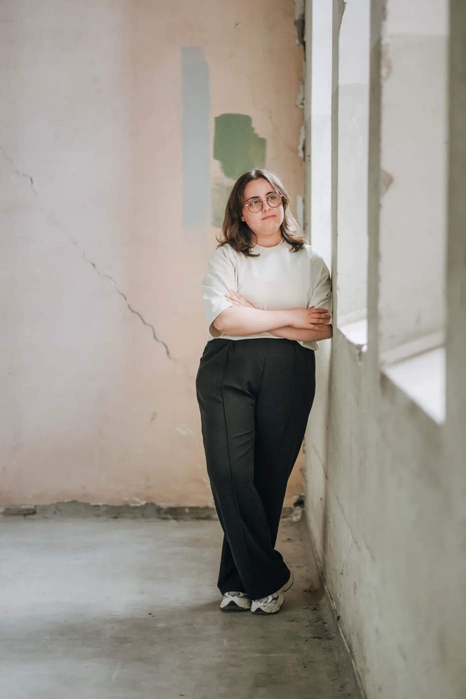 Eine Person lehnt mit verschränkten Armen an der Wand, schaut nachdenklich aus dem Fenster in einem kargen Raum.