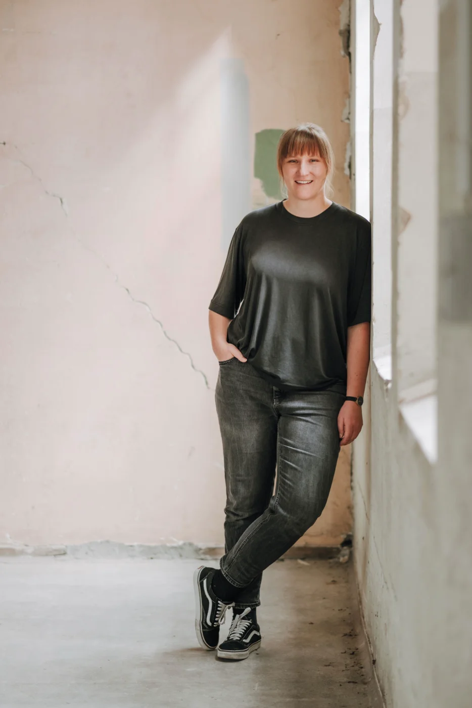 Person in schwarzem T-Shirt und Jeans lehnt lächelnd an einer Wand in einem hellen Raum.