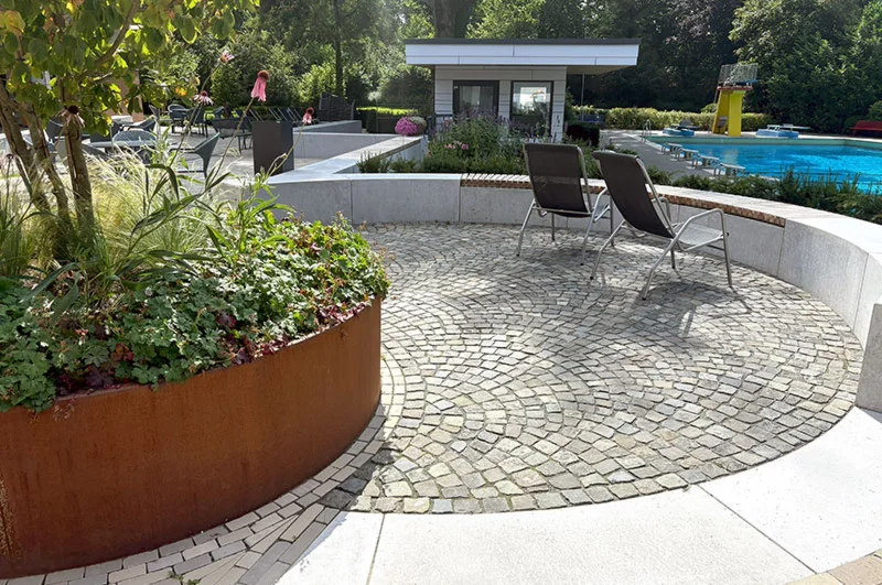 Rundbeet und zwei Liegestühle auf gepflastertem Poolbereich, Schwimmbad und Gebäude im Hintergrund, sommerliche Atmosphäre.