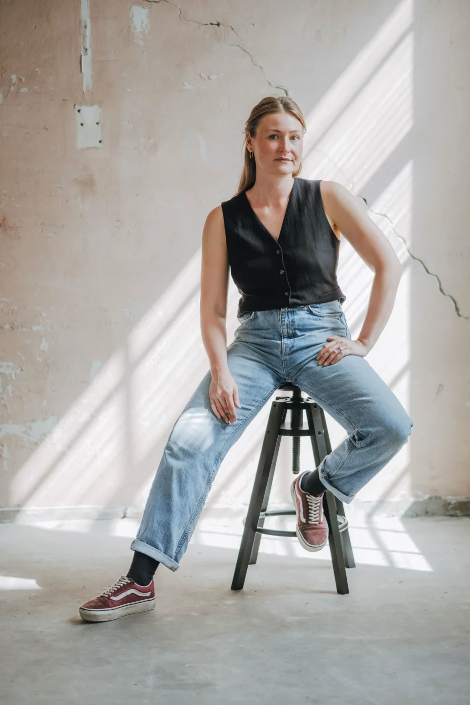 Frau in schwarzer Bluse und Jeans sitzt auf Barhocker in sonnigem Raum, Backsteinwand im Hintergrund.