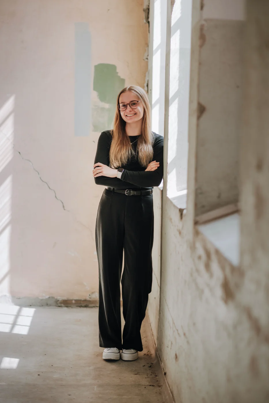 Frau mit langen Haaren und Brille lehnt lächelnd an einer Wand in einem lichtdurchfluteten Raum.