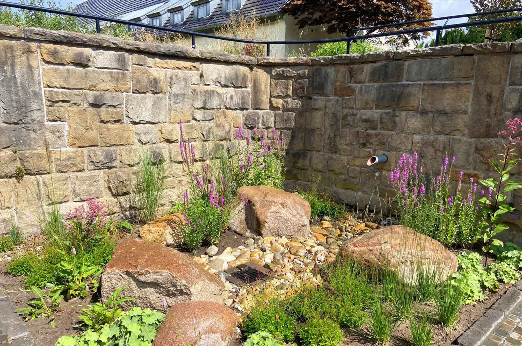 Steingarten mit bunten Blumen und großen Felsen vor einer Natursteinmauer, im Hintergrund ein Gebäude.