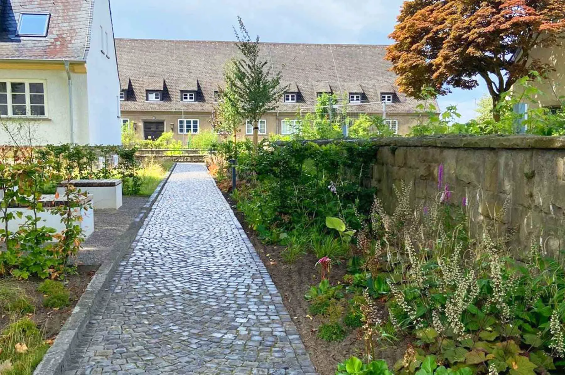 Pflasterweg zwischen Gärten führt zu einem Gebäude mit Satteldach, umgeben von grüner Vegetation und Bäumen.
