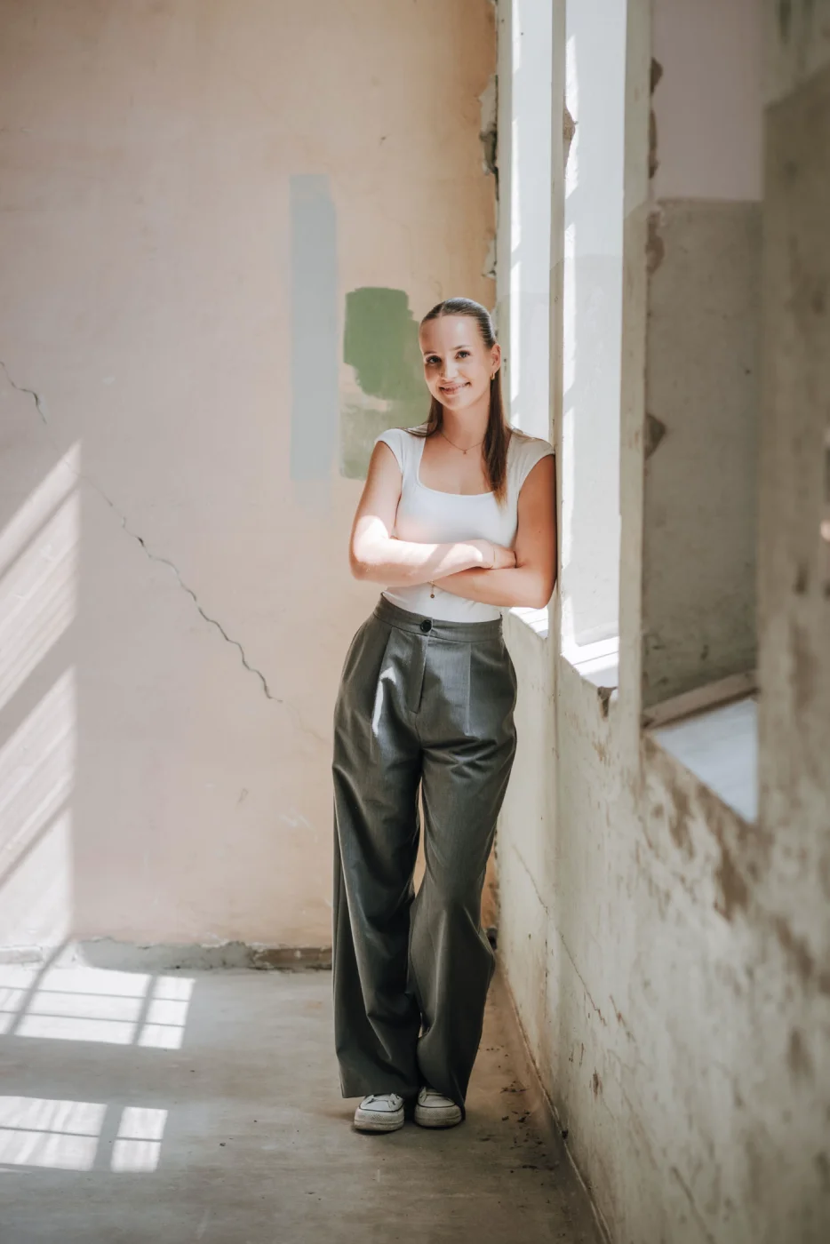 Frau in weißem Top und grauer Hose lehnt lächelnd an einer sonnendurchfluteten Wand in einem verlassenen Raum.
