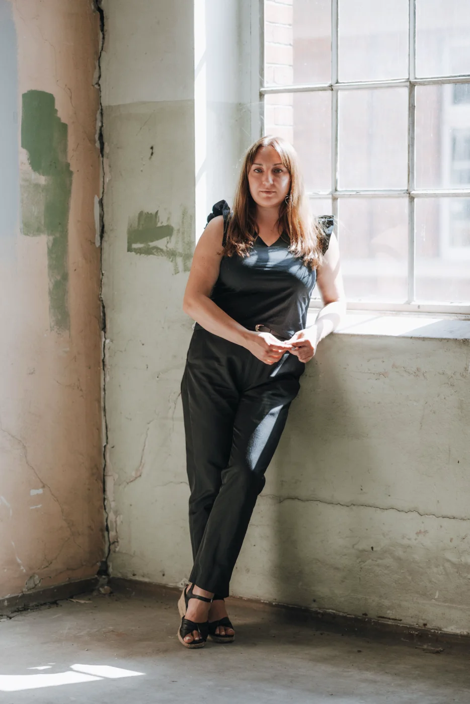 Frau in schwarzem Outfit lehnt an einer Wand neben einem Fenster; Innenraum mit rauem Putz, Sonnenlicht fällt herein.