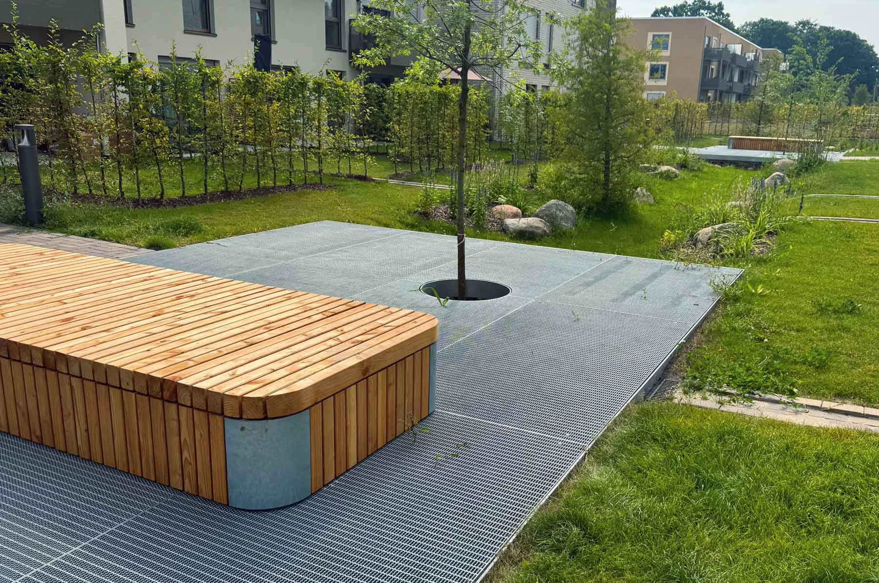 Holzbank auf einem Gitterpodest vor Wohngebäuden, umgeben von grünen Pflanzen und Bäumen.