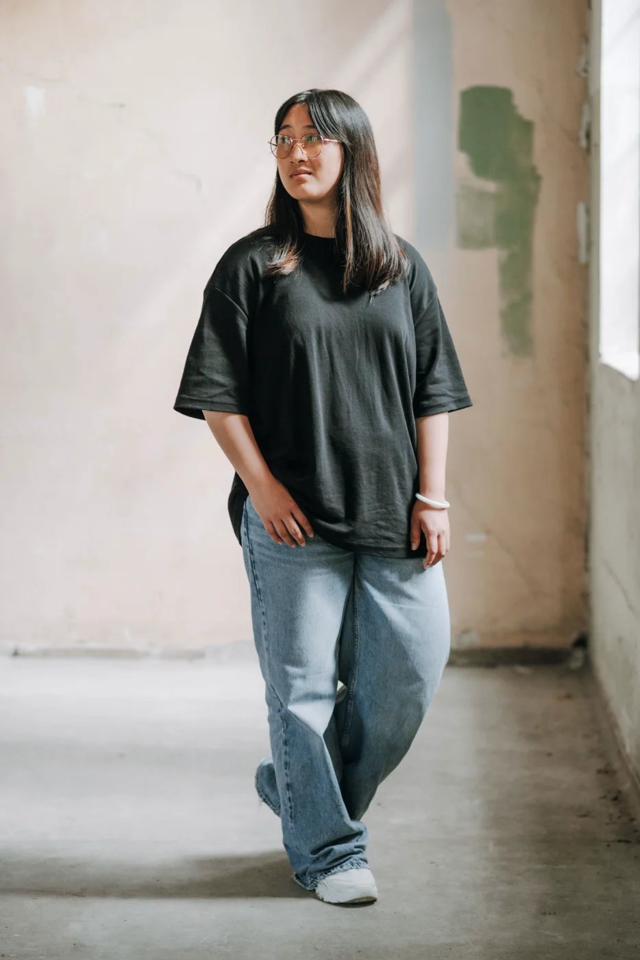 Frau mit Brille, schwarzem T-Shirt und blauer Jeans steht in einem leeren Raum mit beigem, leicht abgenutztem Hintergrund.
